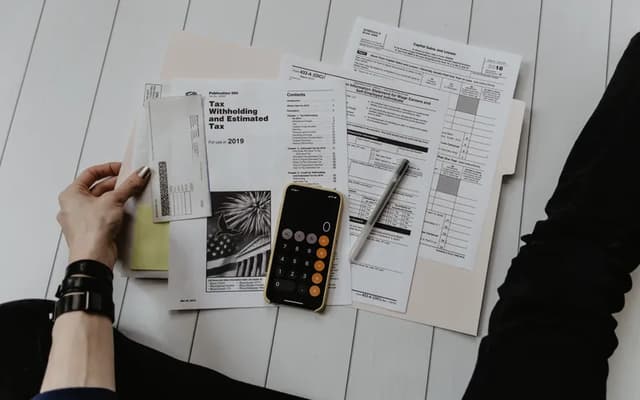Hands sorting a pile of forms and receipts on a desk, next to a phone calculator