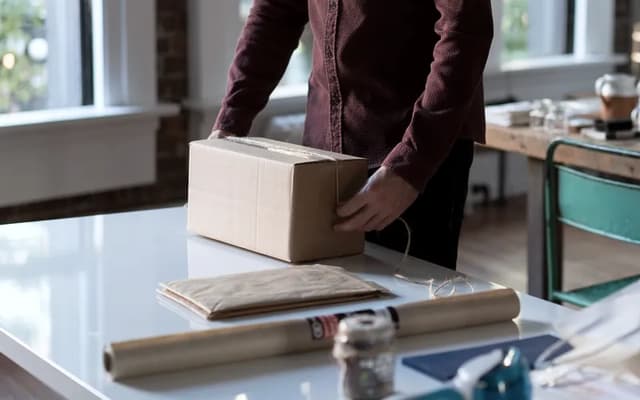 Someone packing an order into a cardboard box at a table by the window